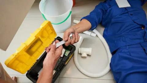 Hands holding a white drain hose and fittings next to a toolbox and a bucket