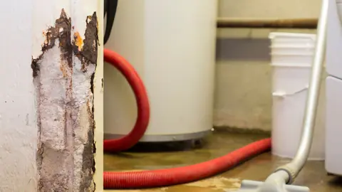 Wet vacuum hose and a bucket beside a water heater with a rusted pipe stand on a wet floor
